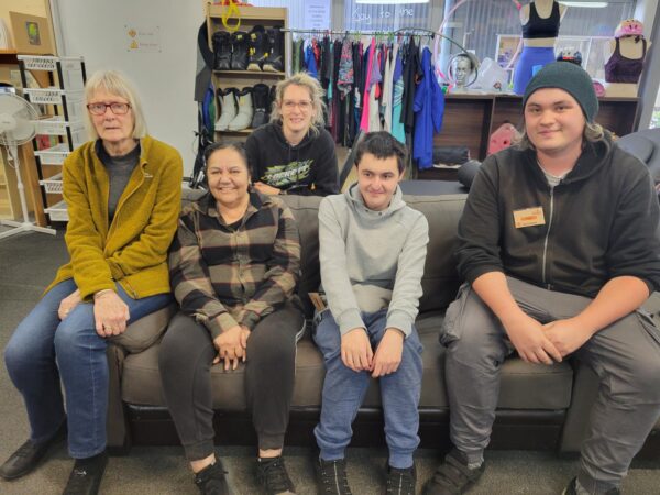 Five Hospice Whanganui volunteers sitting together on a sofa at the Whanganui East Hospice Shop, smiling and relaxed.