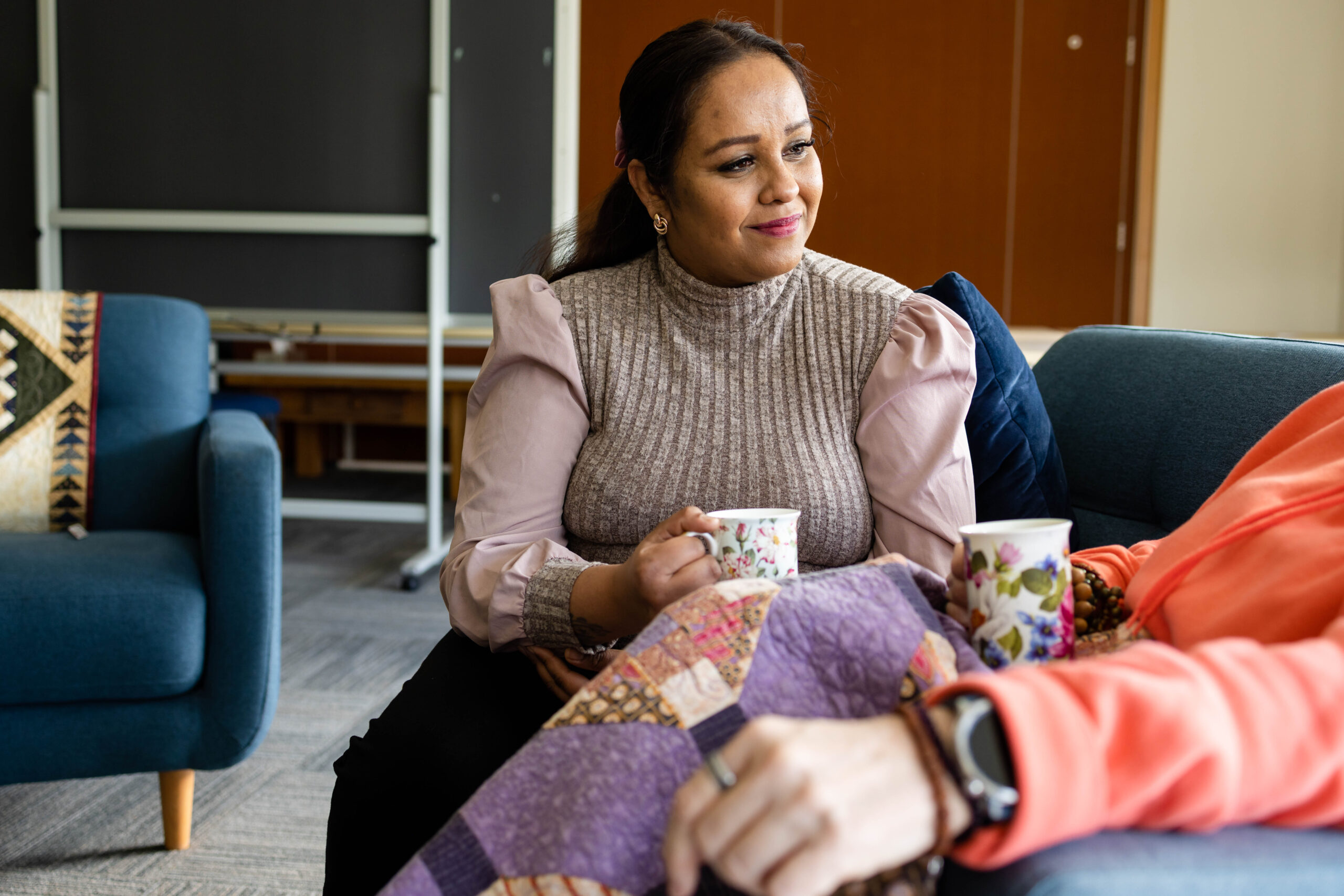 A Hospice Whanganui staff member sits beside a patient, holding a cup of tea and listening with warmth and compassion. The two sit together on a couch, sharing a quiet moment of support.