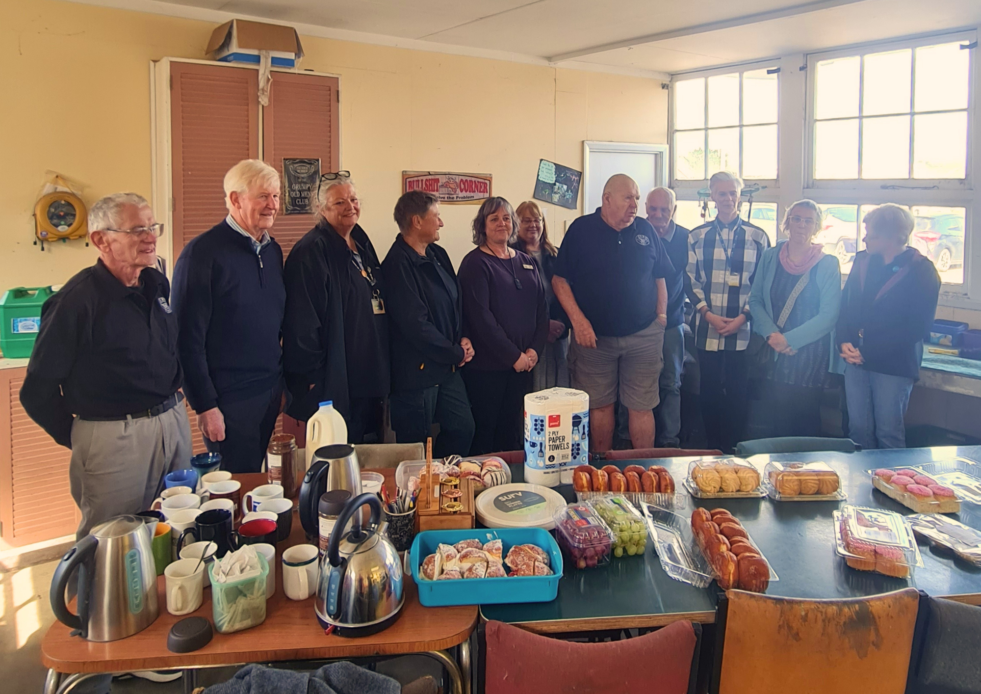 G.O.M.E. members, Hospice Whanganui staff and other local organisation representatives standing together behind a morning-tea table filled with baked goods and hot drinks.