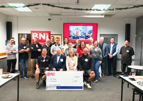 Members of BNI Whanganui stand with Hospice Whanganui CEO Helen Leslie while presenting a donation cheque in a meeting room.