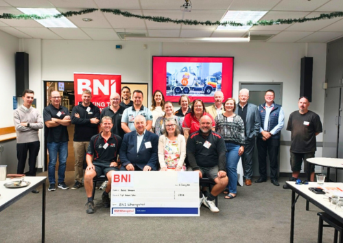 Members of BNI Whanganui stand with Hospice Whanganui CEO Helen Leslie while presenting a donation cheque in a meeting room.